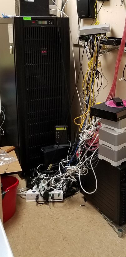 A tower on the floor beside a UPS with unmanaged cabling looped between them